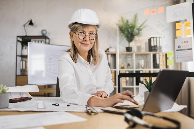 Architetto femminile maturo che utilizza il computer portatile che lavora agli schizzi del piano di costruzione