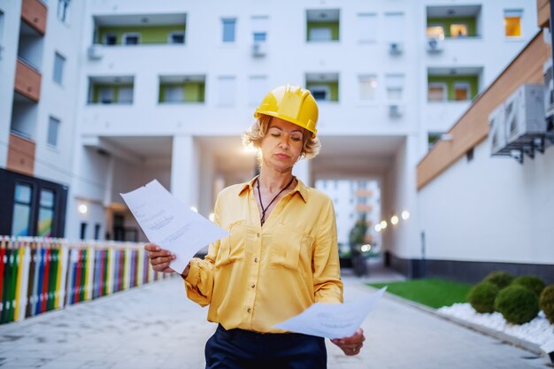 Architetto femminile senior caucasico riuscito con il casco sulla testa che esamina i documenti mentre levandosi in piedi all'aperto.