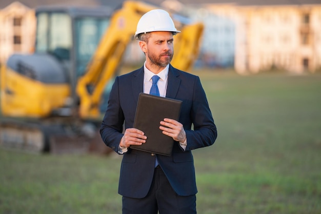 Architetto in un cantiere. Architetto uomo in casco e tuta nella costruzione di edifici domestici moderni. Architetto con giubbotto e tuta di sicurezza. Architetto sicuro in piedi sullo sfondo della casa.