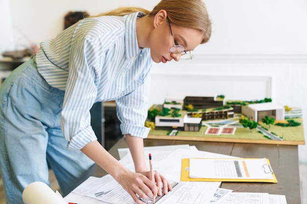 Architetto serio della donna bionda in occhiali che lavora con i disegni mentre progetta il progetto sul posto di lavoro