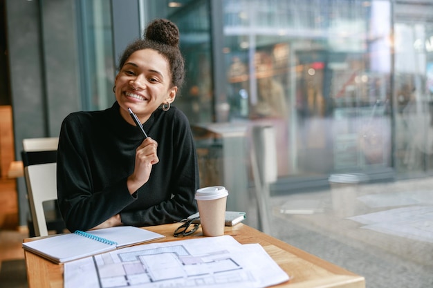 Architetto sorridente della donna che guarda l'obbiettivo mentre lavora nel concetto del lavoro a distanza del caffè