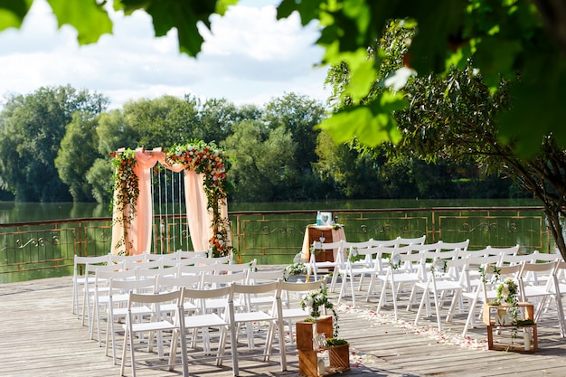 Area della cerimonia di nozze vicino al fiume sul molo. Arco rettangolare in legno, sedie bianche decorate con fiori, verde, petali, foglie e tulle color pesca. Decorazioni rustiche carine e di tendenza