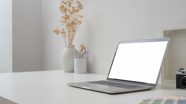 Area di lavoro con laptop a schermo vuoto, pennello, campione di colore, cornice e vaso di ceramica sulla scrivania