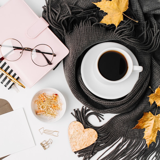 Area di lavoro con taccuino con carta vuota, tazza di caffè avvolta in sciarpa, occhiali. Elegante scrivania da ufficio. Autunno o inverno concetto. Disposizione piatta, vista dall'alto