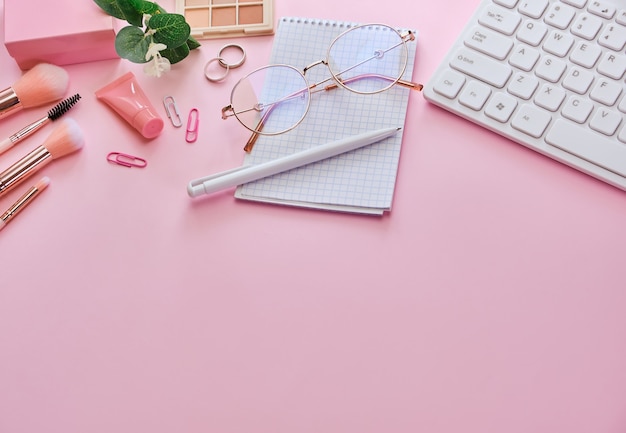 Area di lavoro con tastiera, blocco note, occhiali, penne, accessori di bellezza sulla superficie rosa