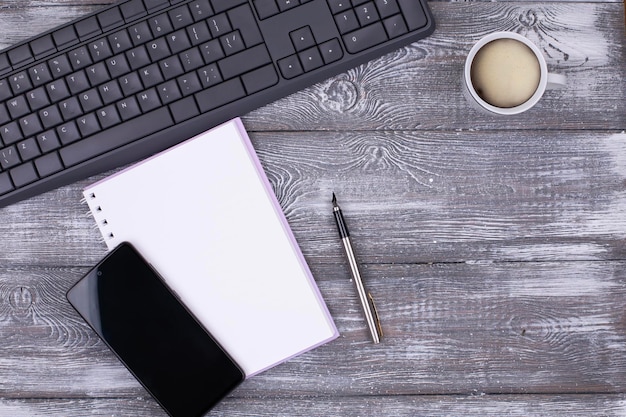 Area di lavoro con tastiera tazza di caffè blocco note a molla vuota per idea nota obiettivo piano concetto di scrittura penna per smartphone su sfondo di legno imbiancato grigio Copia spazio piatto lay