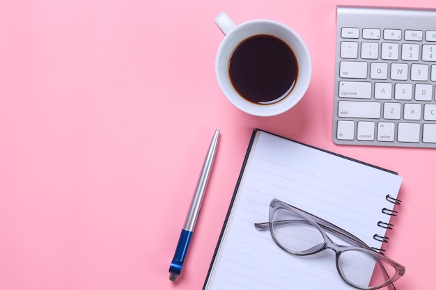 Area di lavoro da scrivania femminile con tastiera, notebook, occhiali, penna e tazza da caffè su sfondo rosa