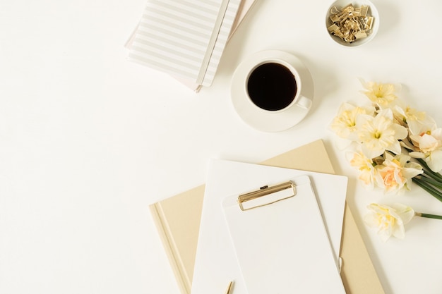 Area di lavoro del tavolo dell'ufficio domestico. Blocco appunti foglio bianco con copia spazio, tazza di caffè, fiori di narciso, cancelleria su bianco