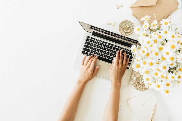 Area di lavoro della scrivania del Ministero degli Interni con il computer portatile, il mazzo dei fiori della margherita della camomilla e il taccuino su superficie bianca