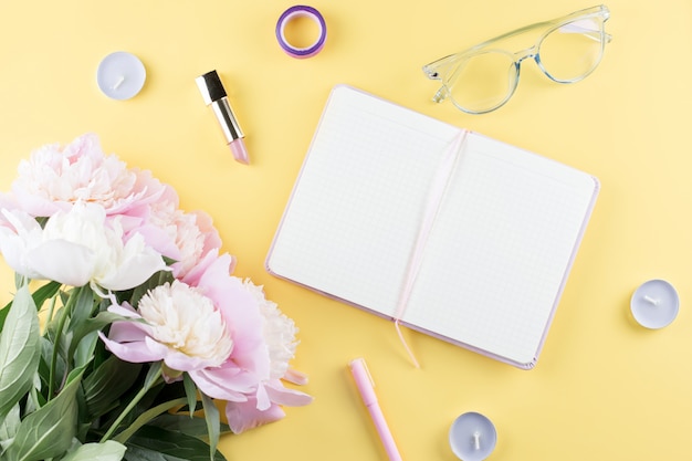 Area di lavoro femminile con taccuino in bianco aperto, fiori di peonia, occhiali da vista