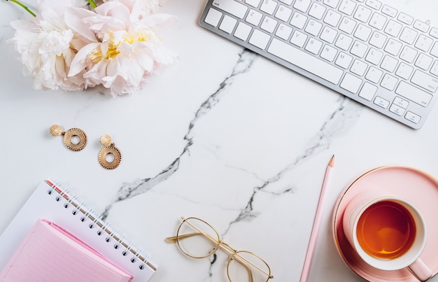 Area di lavoro femminile dell'ufficio domestico con una tazza di tè e fiori di peonia su sfondo di marmo bianco vista dall'alto f...