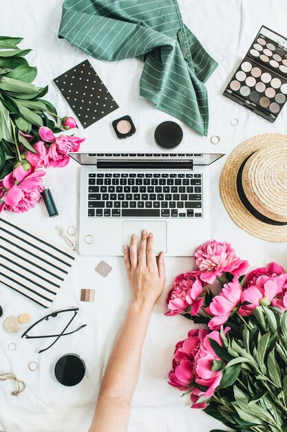 Area di lavoro femminile della scrivania da ufficio piatta e vista dall'alto con laptop, fiori di peonia, occhiali, cappello di paglia, cosmetici, accessori. Donna che lavora al computer.