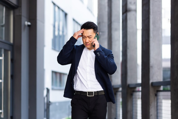 Arrabbiato e serio uomo d'affari asiatico di successo spiega le informazioni ai dipendenti usando il telefono, parla vicino all'ufficio fuori