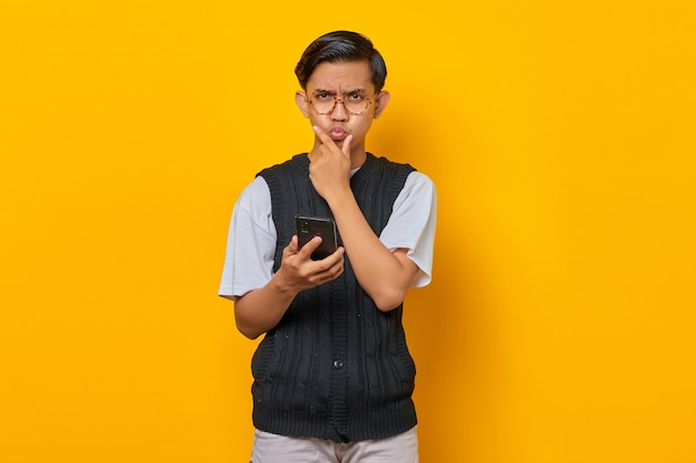 Arrabbiato giovane asiatico con il dito sul mento e tenendo il telefono cellulare su sfondo giallo