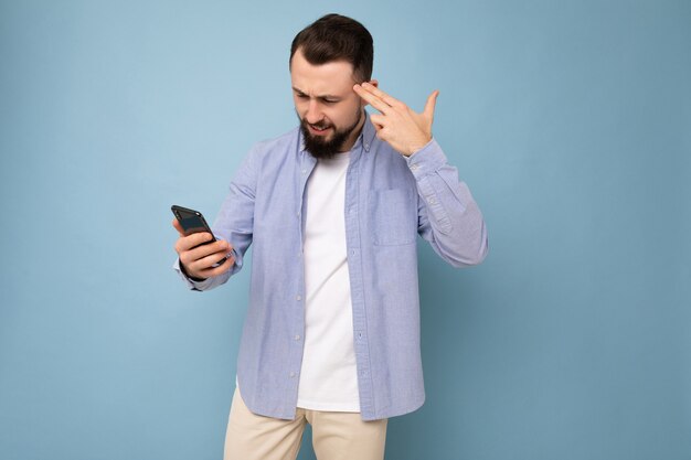 Arrabbiato sconvolto bel giovane brunetta con la barba lunga uomo con barba che indossa elegante t-shirt bianca e maglietta blu isolate su sfondo blu con spazio vuoto tenendo in mano e utilizzando il telefono leggendo le notizie onl