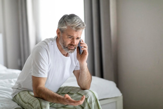 Arrabbiato uomo di mezza età seduto sul letto ha una conversazione telefonica