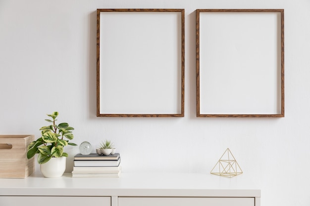 Arredamento minimalista per la casa degli interni con due cornici per foto in legno marrone sullo scaffale bianco con libri, bella pianta in vaso elegante e accessori per la casa. Muro bianco.