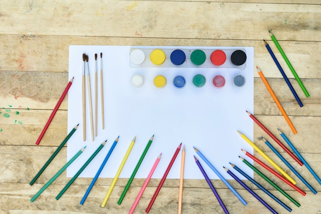 Art. Molte matite colorate, pennelli e vasetti di vernice su un foglio di carta bianco. Tavolo di legno