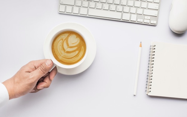 Arte del latte della tazza di caffè della tenuta della mano maschio sul tavolo da lavoro moderno bianco