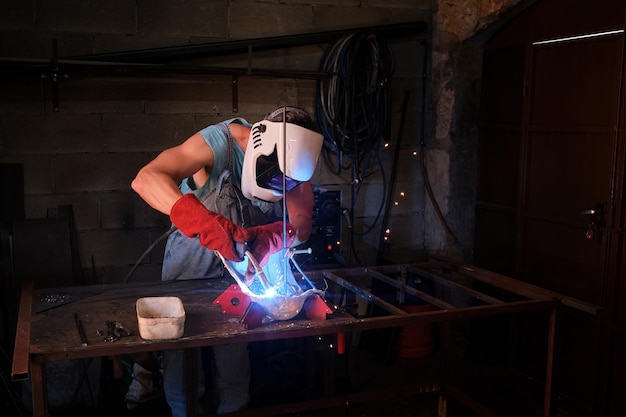 Artigiano maschio irriconoscibile con casco e guanti in piedi al banco di lavoro e saldando dettagli metallici in forma di gesso usando una torcia mentre lavora in un laboratorio professionale