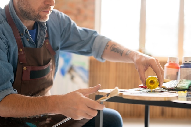 Artista contemporaneo che versa tempera gialla liquida sulla tavolozza mentre è seduto al tavolo in studio e va a lavorare su un nuovo dipinto