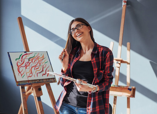 Artista dai capelli scuri che tiene in mano un pennello e disegna un quadro su tela.