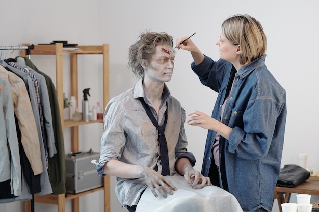 Artista del viso femminile contemporaneo con pennello che applica cerone zombie sulla fronte di un giovane uomo d'affari o attore in ufficio o in studio