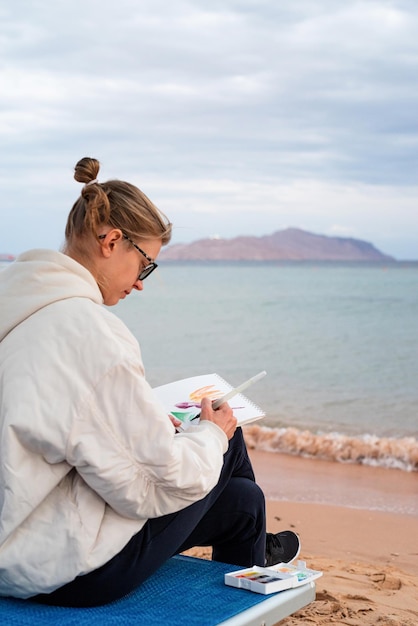 Artista della giovane donna che dipinge o fa schizzi di viaggio usando l'acquerello in riva al mare