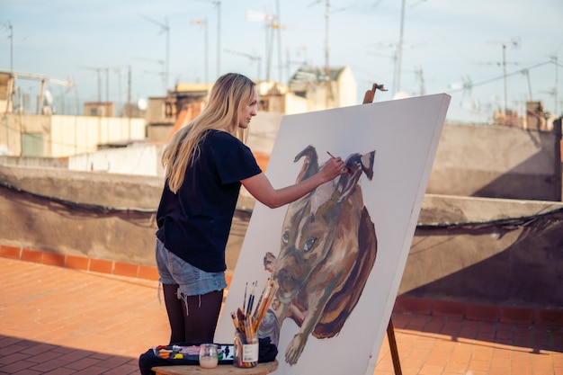 Artista della pittura della giovane donna che disegna a casa il tetto. Immagine di bulldog su grande tela. Arte all'aperto