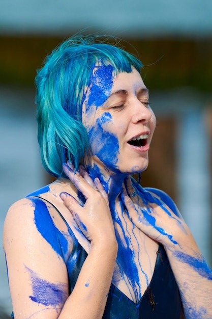 Artista di performance artistica donna dai capelli blu in abito blu scuro imbrattato con pittura a guazzo indaco sul suo corpo sulla spiaggia, primi piani. Body painting creativo, concetto di body art, performance art all'aperto