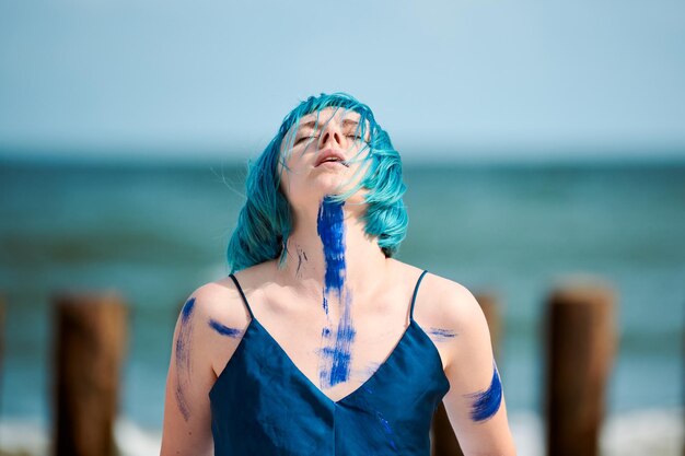 Artista di performance artistica donna dai capelli blu in abito blu scuro imbrattato con pitture a guazzo blu che ballano sulla spiaggia, primo piano. Body painting e body art concept, performance art espressiva all'aperto