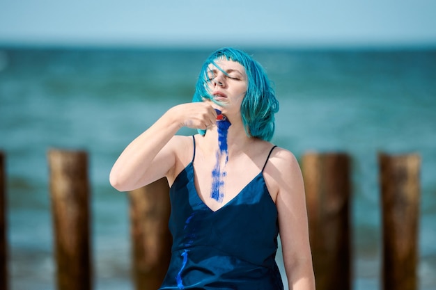 Artista di performance artistica donna dai capelli blu in abito blu scuro spalmato di pittura a guazzo indaco sul suo corpo con spazzole sulla spiaggia. Body painting creativo, concetto di body art, performance all'aperto