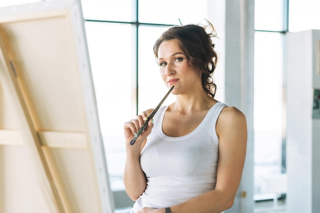 Artista donna pensante in abiti casual bianchi con pennelli davanti al cavalletto nella galleria d'arte