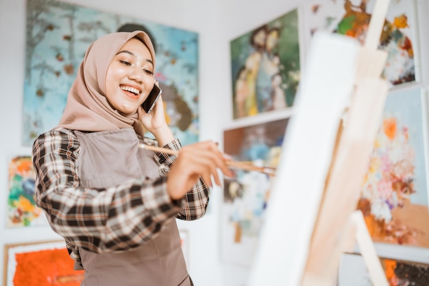 Artista femminile asiatico musulmano che dipinge su tela e parla al telefono