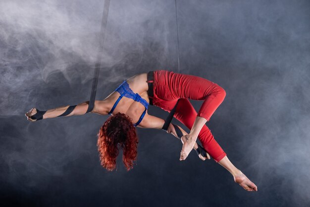 Artista femminile del circo aereo atletico, sexy e flessibile con la rossa su cinghie aeree sul nero.