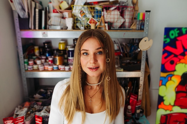 artista femminile sorridente in abiti casual in piedi in officina sullo sfondo degli scaffali