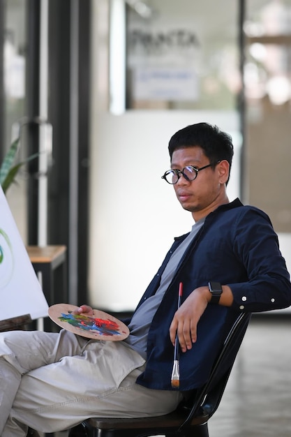 Artista uomo asiatico ritratto seduto davanti a tela e guardando la fotocamera