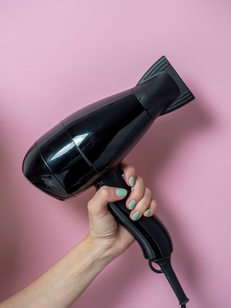 Asciugacapelli nero in mano di una donna su uno sfondo rosa. Prodotto per la cura dei capelli. Foto verticale