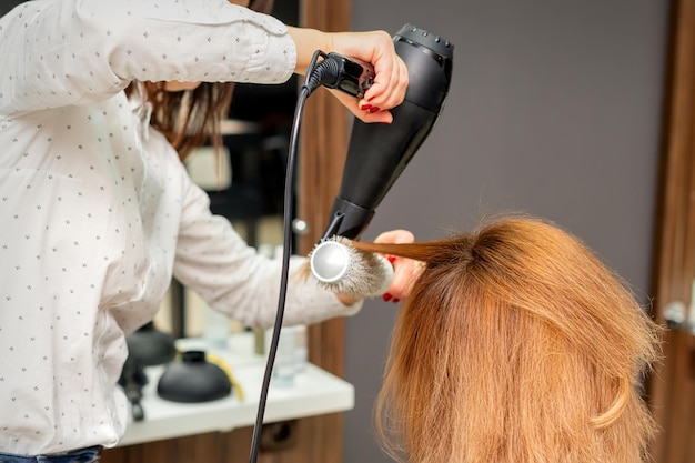 Asciugare i capelli nello studio dei capelli. Lo stilista femminile del parrucchiere asciuga i capelli con un asciugacapelli e capelli rossi della spazzola rotonda di una donna in un salone di bellezza.