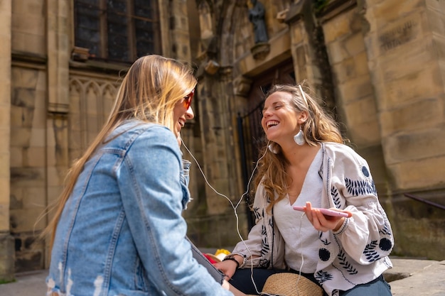 Ascoltando musica in città due amiche caucasiche che sorridono e condividono le cuffie e ascoltano musica in streaming