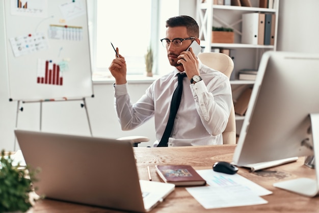 Ascolto delle esigenze dei clienti. Giovane serio in abiti da cerimonia che parla al telefono e gesticola mentre è seduto in ufficio