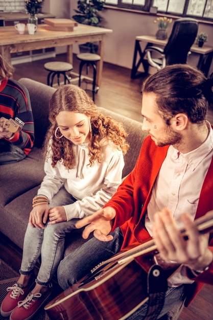 Ascolto fratello. Ragazza riccia piacevole che indossa una felpa con cappuccio bianca che ascolta il fratello che suona la chitarra