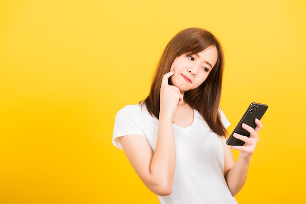 Asian felice ritratto bella giovane donna carina adolescente in piedi indossare t-shirt lei utilizzando tenendo il telefono cellulare intelligente e pensando guardando al telefono isolato, studio girato sfondo giallo con copia spac