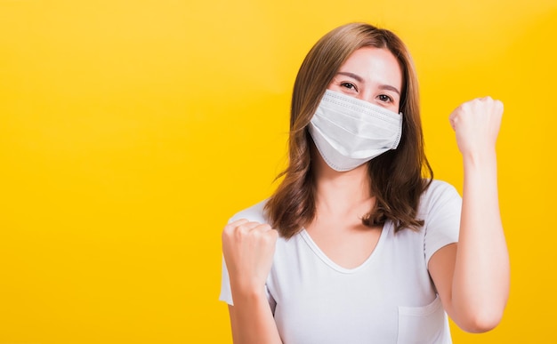 Asiatica bella giovane donna felice che indossa maschera facciale protezione filtro polvere pm2.5, virus COVID e inquinamento atmosferico alza le mani felice eccitato allegro dopo essersi ripreso dalla malattia su sfondo giallo