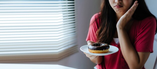 asiatica giovane bella donna sensazione di dolore da mal di denti e tenendo deliziosa ciambella glassata al cioccolato sul piatto