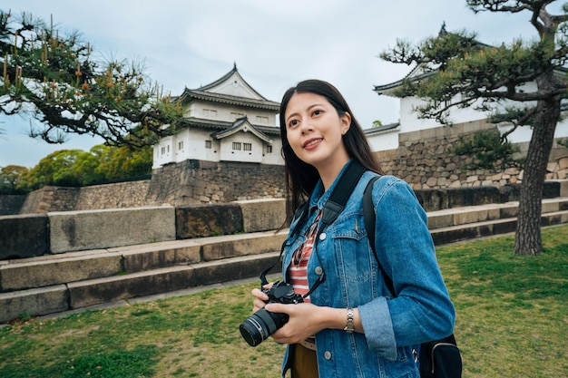 asiatico giovane ragazza turista tenendo la fotocamera nelle mani in piedi nel parco verde con il castello di osaka in background. Ritratto di stile di vita estivo all'aperto di bella donna che si diverte nel viaggio in jp.