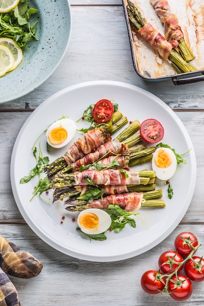 Asparagi freschi avvolti in pancetta su un piatto bianco con pomodori rucola e uova vista dall'alto.