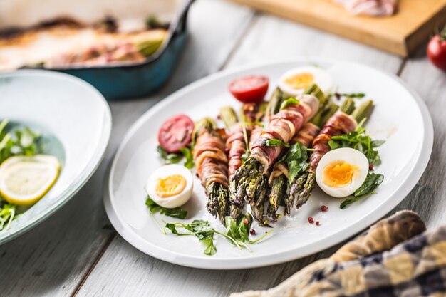 Asparagi freschi avvolti in pancetta su un piatto bianco con pomodorini e uova di rucola.