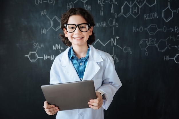 Aspettando il mio primo paziente. Piccolo dottore capace ottimista in piedi vicino alla lavagna in laboratorio mentre si gode la lezione di medicina e utilizza il gadget