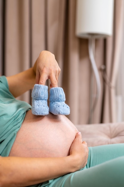 Aspettando la madre seduta sul divano e tenendo le dita in minuscoli stivaletti blu Calzini per un bambino su una grande pancia incinta Tende sfocate e paralume sullo sfondo